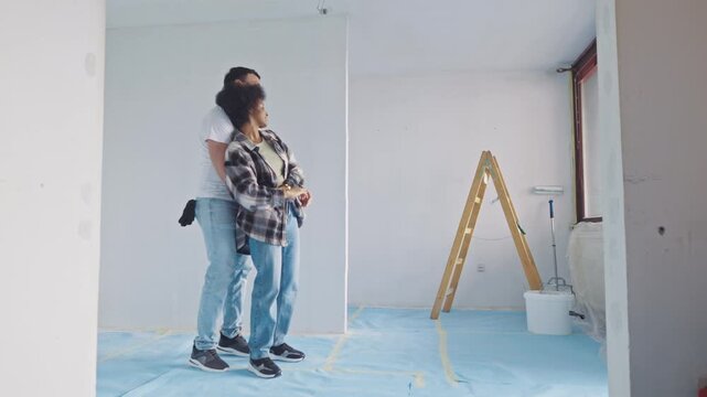 In a sunlit room under renovation, a young woman and a man engage in playful interaction, surrounded by blue drop cloths and a ladder, hinting at ongoing home improvement.