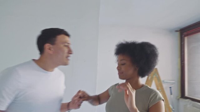 In a sunlit, partially renovated apartment, a joyful couple dances energetically, celebrating their home improvement progress. The man in a white t-shirt and the woman in a green top laugh and twirl.