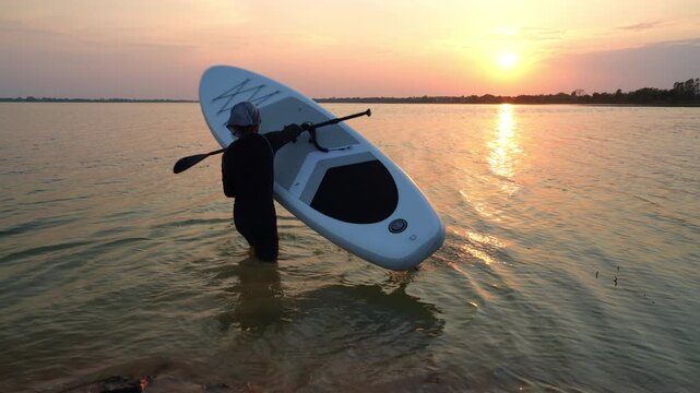 The silhouette of a person carrying a paddle board and playing at sunset symbolizes outdoor exercise, water sports, summer activities, a healthy lifestyle, and adventure travel.