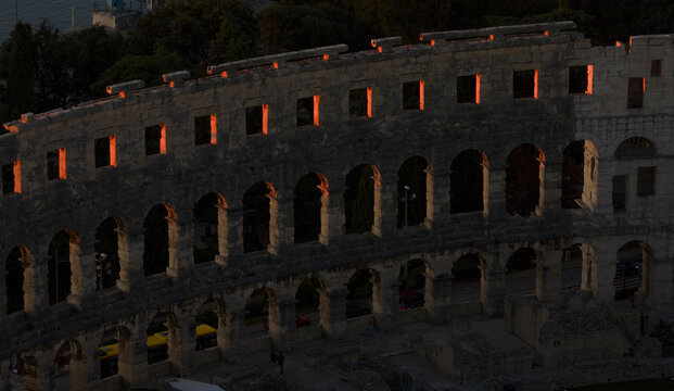 Aerial view of the ancient Roman amphitheater, bathed in the warm glow of the setting sun, casting long shadows on its weathered stone facade, Pula, Istria, Croatia.