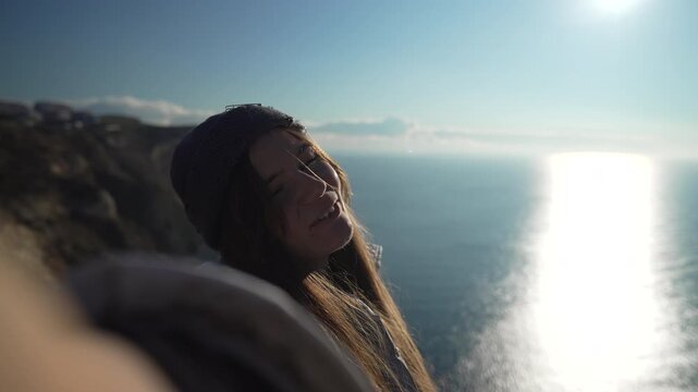 Woman selfie travel, happy young female takes video standing on sunny cliff overlooking vast blue ocean view.