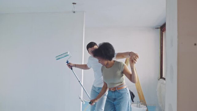 A diverse couple dances playfully in a bright, minimalist room during a home renovation, using a paint roller and brush. The modern, white-walled space, equipped with a stepladder and paint supplies.