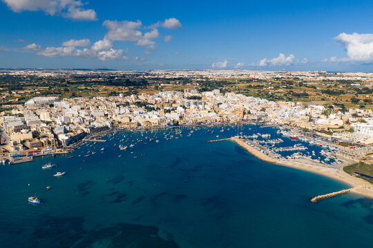 Aerial view of the harbor dotted with colorful boats against the backdrop of the town's white buildings and the azure sea, Marsaxlokk, Malta.