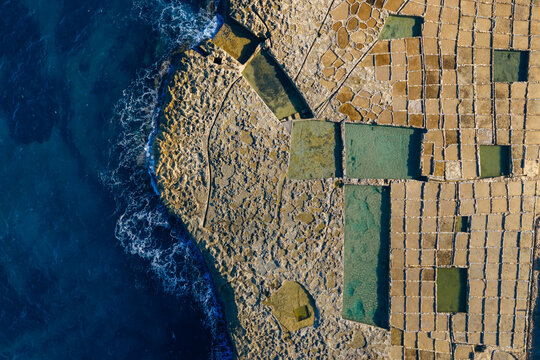 Aerial view of geometric salt pans meet the dark blue sea, creating a stunning contrast of textures and colors, Zebbug, Malta.