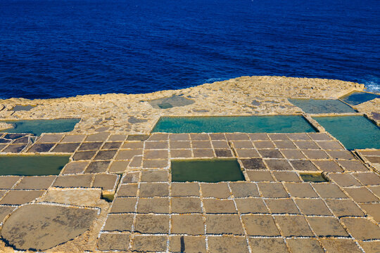 Aerial view of geometric salt pans reflecting the azure sky meet the rugged coastline, creating a mosaic of textures and tones, Zebbug, Malta.