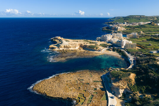 Aerial view of the rugged coastline meeting the turquoise waters, a tranquil bay nestled beside stone buildings and green fields, Zebbug, Malta.