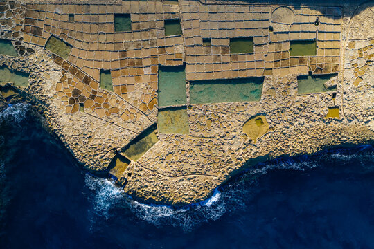 Aerial view of the ancient salt pans carved into the rugged coastline, where turquoise waters meet sun-baked earth in a mosaic of textures and tones, Zebbug, Malta.