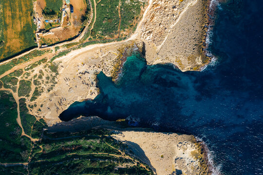 Aerial view of the rugged coastline where turquoise waters meet the tan rocks and green vegetation, St.Peter's Pool, Marsaxlokk, Malta.