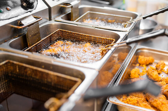 Industrial deep fryers cooking breaded chicken in hot oil in a commercial kitchen, featuring wire baskets and stainless steel equipment in a shallow depth of field