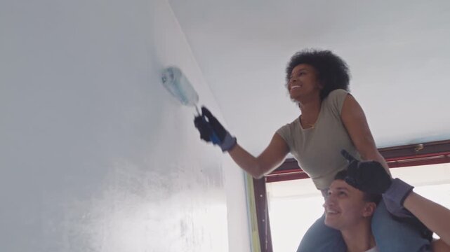 A diverse couple collaborates on home renovation, with a woman spreading plaster on a wall and a man supporting her from below in a bright apartment setting.
