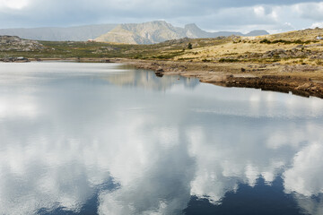 Fototapeta premium A tranquil glacial lake sits in a rocky mountain basin with dark, dramatic storm clouds gathering over the distant peaks. Stock photo