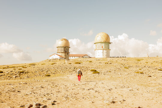 Person hiking towards old radar domes and buildings situated on a high, barren mountain plateau under a blue sky. Stock photo