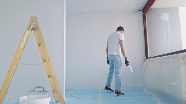 A young man, focused and engaged in renovating his apartment, carefully applies plaster to the wall. Dressed in casual attire and surrounded by tools, he sits amid a makeshift workspace.