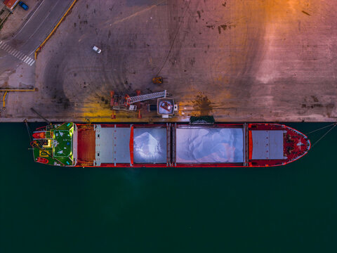 Aerial view of a cargo ship docked at the port, its red hull contrasting with the deep green water and the pale dockside, Vasto, Abruzzo, Italy.