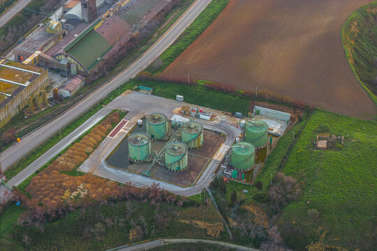 Aerial view of industrial tanks gleaming against the backdrop of cultivated fields and a winding road, creating a stark contrast between human activity and nature, Vasto, Abruzzo, Italy.