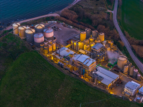 Aerial view of the golden-lit industrial plant casting a warm glow against the fading twilight, nestled between the sea and verdant slopes, Vasto, Abruzzo, Italy.