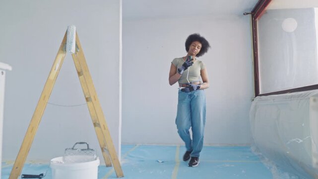 A young woman with curly hair dances joyfully in her brightly lit, renovation-filled apartment, holding paint rollers and surrounded by ladders and protective coverings.