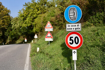 Obraz premium Collection of roadside traffic signs along a rural road in the Province of Siena, Italy, including a 50 km/h speed limit, snow chain requirement notice, and warning signs ahead.