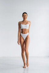 Slim female model wearing white bikini poses confidently in a bright studio with a clean, minimalistic background emphasizing her physique and fashion style