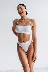 Fototapeta premium Slim female model wearing white lingerie poses confidently against a plain background, showcasing a fit physique and contemporary fashion style in a studio setting