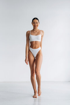 Slim female model wearing white bikini poses confidently in a bright studio with a clean, minimalistic background emphasizing her physique and fashion style