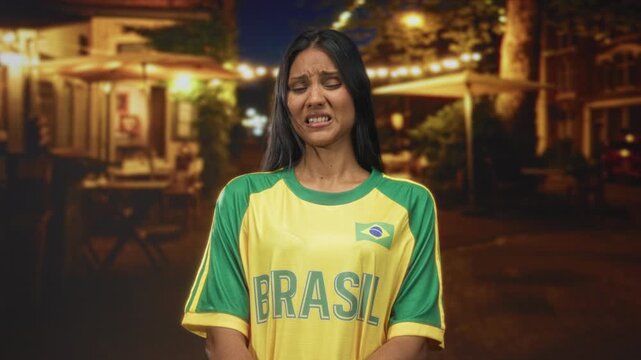 Young brunette woman in yellow brazil jersey grimacing, face contorted under cafe lights on street; unease.