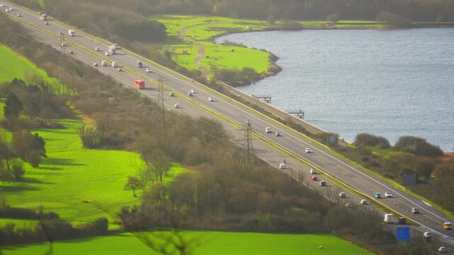 Cars Moving Along M4 Motorway in Wales with Nearby Boating Lake with Lush Green Fields. Illustration of Modern Highway with Natural Surroundings. 4K Clip.