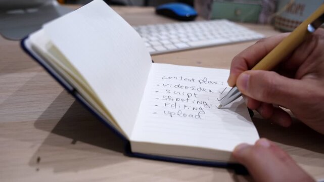 Close-up of a hand marking a content plan checklist in a notebook on a wooden office desk, with pen and blurred keyboard, showing video idea, script, shooting, editing, and upload tasks.