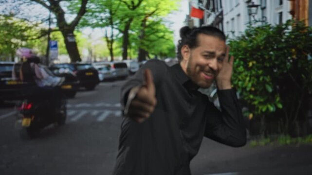 Man giving thumbs up with hand to ear on city street near parked cars and leafy trees, closeup on thumb and smile; playful confidence.