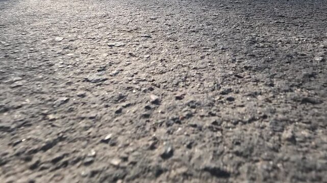 Extreme close view of rough asphalt pavement as the camera moves smoothly above the road surface, revealing sharp stone texture, shifting light, and soft shadow drift in bright outdoor sun.