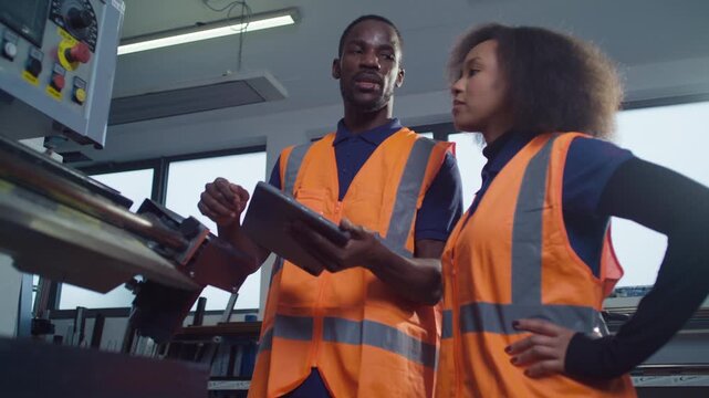 Two labour workers in industrial plant, looking at a tablet, discussing production and digital technology integration in factory operations.