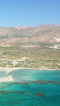 Scenic aerial shot of Elafonisi beach with clear turquoise waters, sandy shoreline, and rugged mountains in Crete, Greece under a bright blue sky.