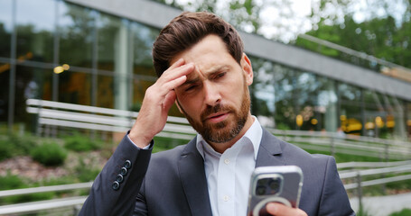 Close-up of a concerned bearded businessman in a business suit, holding a smartphone and reacting...