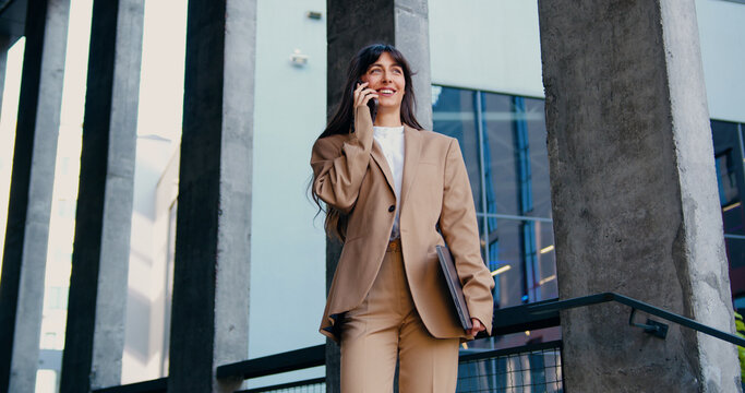 Confident businesswoman in a formal suit holding a laptop and talking on the phone while walking outdoors. Modern glass office building in the background. Business communication, important call