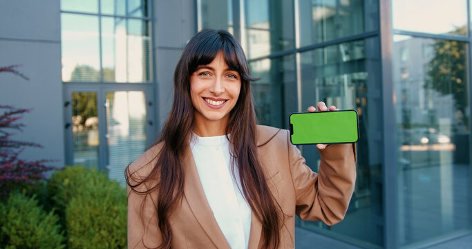 Confident businesswoman in suit smiling and holding a smartphone with a green screen. Perfect for showcasing mobile apps, user interface and UX designs, software demos, or tech startup presentations.