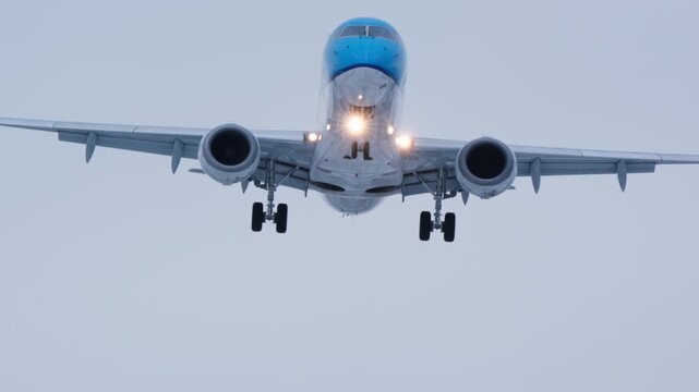 A commercial airplane with landing gear down and lights on approaches the camera for landing, getting closer before exiting the frame. Overcast sky background.