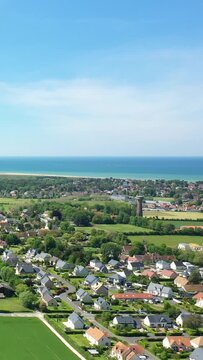 Scenic aerial shot of Merville-Franceville village with green fields, residential houses, and the coastline of Normandy, France under a clear blue sky.