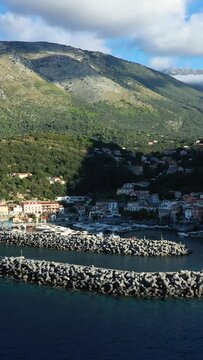 Scenic view of Maratea harbor with yachts, breakwaters, and lush green mountains in southern Italy under a partly cloudy sky.