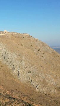 Wide view of the Acrocorinth fortress atop a rugged, sunlit hill under a clear blue sky in Greece. The historic stone walls are visible on the summit, surrounded by dry, rocky terrain.