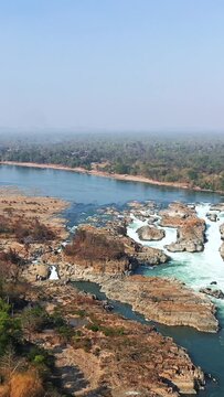 Drone shot of Khone Falls with cascading water over rocky terrain and lush forest along the riverbank in Laos. Clear blue sky and expansive natural scenery create a dramatic landscape.