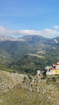 Scenic view of a small hilltop village surrounded by lush green mountains under a bright blue sky in Aieta, Italy. Picturesque rural landscape with historic stone buildings and rugged terrain.
