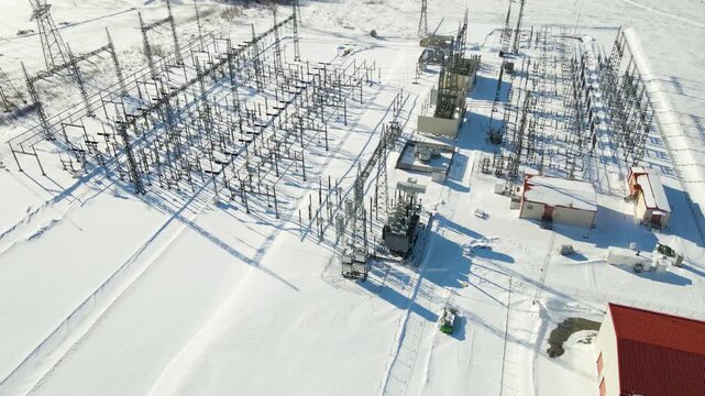 Aerial approach toward a large electrical substation with transformers and pylons casting long shadows across snowy terrain powering modern digital infrastructure in a cold winter environment.