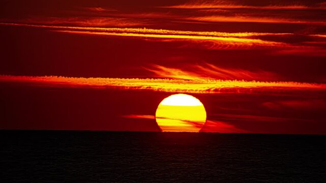 A full yellow sun rises in an orange sky over the still dark ocean water.