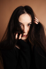Fototapeta premium Elegant young woman with long wavy dark hair posing in a professional photo studio on a solid brown background, sophisticated portrait, beauty and fashion concept, Prague, March 2025