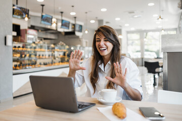 Fototapeta premium A cheerful young woman waves and laughs while having a video call on her laptop at a bakery table with coffee and a croissant.