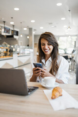 A happy young woman checks her smartphone while sitting at a table with a laptop, coffee, and...