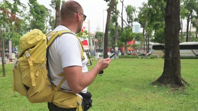 White Traveler With Yellow Backpack Checking Smartphone In Green City Park, Trees And Buses In Background, Sunglasses, Casual Gear, Contemplative Pause While Navigating Saigon Streets