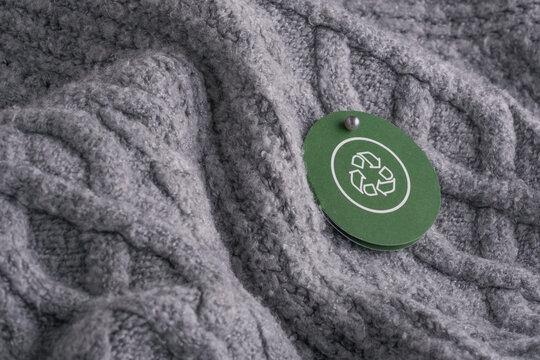 A circular green tag with a recycling symbol hangs from on gray knitted fabric. Concept of sustainable development, textile recycling, and conscious consumption in fashion.