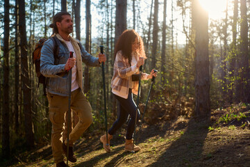 Fototapeta premium Active man and woman enjoying an outdoor adventure, walking uphill on a forest path with hiking poles, bathed in warm morning light filtering through the trees
