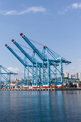 Fototapeta premium A row of large blue container cranes operating at a busy industrial port. Stacked shipping containers and waterfront structures are visible around the cranes, with an urban cityscape in the background